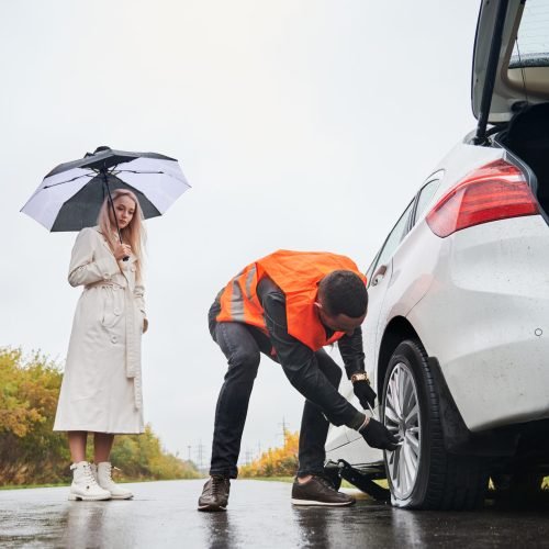 Male auto mechanic unscrewing lug nuts on car wheel while elegant woman holding umbrella. Young man in vest repairing woman automobile on the road. Concept of emergency road service.